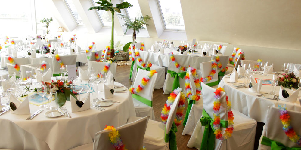 Dekoration für einen Geburtstag im Schokoladenmuseum. Thema Hawaii. Exotische Blumen Gestecke auf den Tischen. Weiße Hussen über die Stühle mit grünen schleifen. Dazu übrige Blumenketten an jedem Stuhl. In den Ecken Palmen und exotische Hawaii Dekoration.