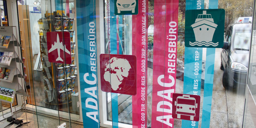 Schaufenstergestaltung Thema Reisebüro. Verschiedene Piktogramme, Schiff, Flugzeug, Auto, Bus und Weltkugel. Dahinter Banner in verschiedenen Breiten in verschiedenen Sprachen für gute Reise. Farbigkeit Brombeer und Petroll.