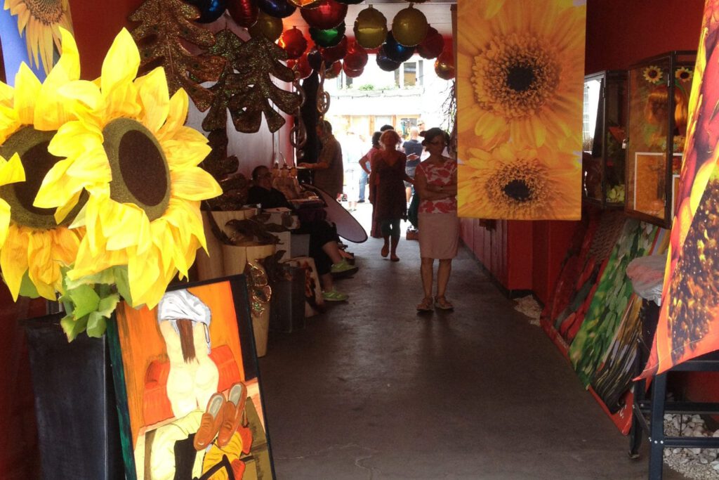 Eindrücke von unserem Hofmarkt in (Flohmarkt) - zu sehen die rote toreinfahrt gesäumt mit großen Banner mit Sonnenblumen und im Vordergrund ein Gemälde mit Akt einer fülligen Frau.